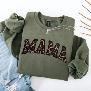 May include: An olive green sweatshirt with the word "MAMA" in leopard print lettering. A silver necklace with a circular pendant rests on the sweatshirt. The sweatshirt is folded on top of a pair of blue jeans.