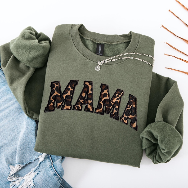 May include: An olive green sweatshirt with the word "MAMA" in leopard print lettering. A silver necklace with a circular pendant rests on the sweatshirt. The sweatshirt is folded on top of a pair of blue jeans.