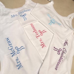 May include: White tank tops with embroidered names and bow designs in various colors. The names "Mrs. Stephens", "Mrs. Murray", "Mrs. Terhune", "Mrs. McGraw", and "Mrs. Christianson" are visible. The tank tops are arranged on a light-colored surface.
