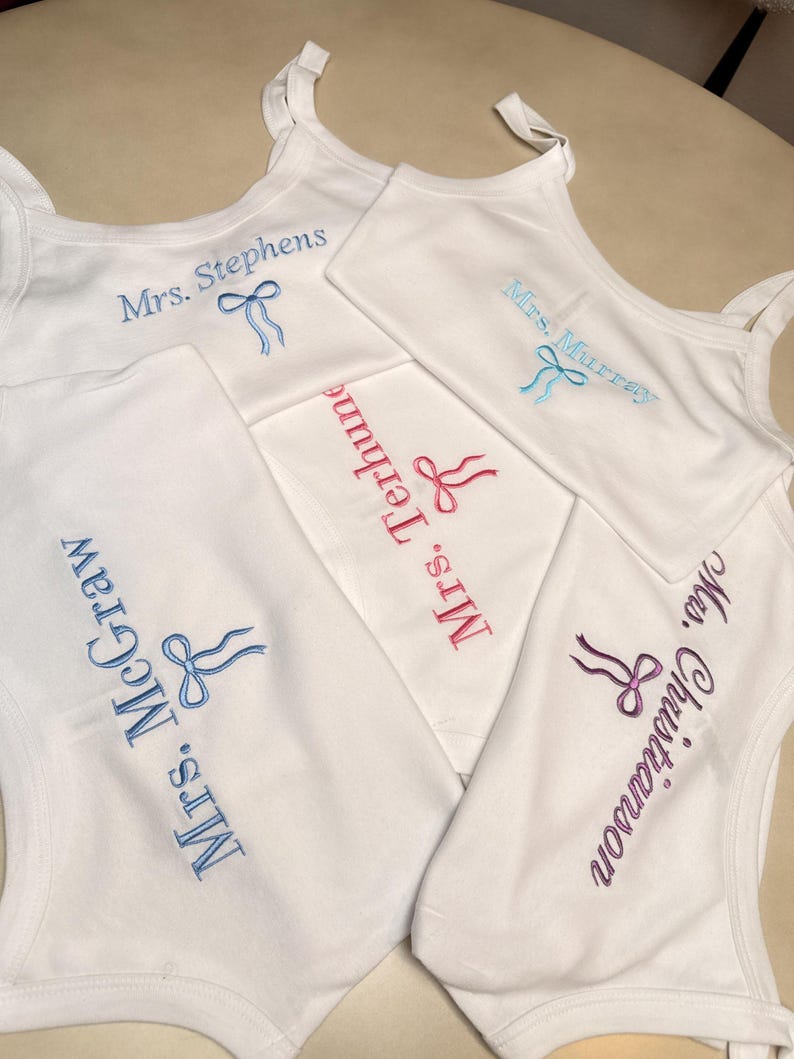May include: White tank tops with embroidered names and bow designs in various colors. The names "Mrs. Stephens", "Mrs. Murray", "Mrs. Terhune", "Mrs. McGraw", and "Mrs. Christianson" are visible. The tank tops are arranged on a light-colored surface.