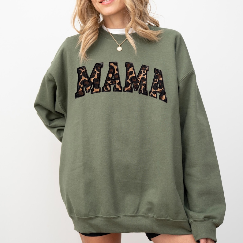 May include: Olive green sweatshirt with the word "MAMA" in a leopard print design across the chest. The sweatshirt has a crew neck and long sleeves. The overall style is casual and comfortable.