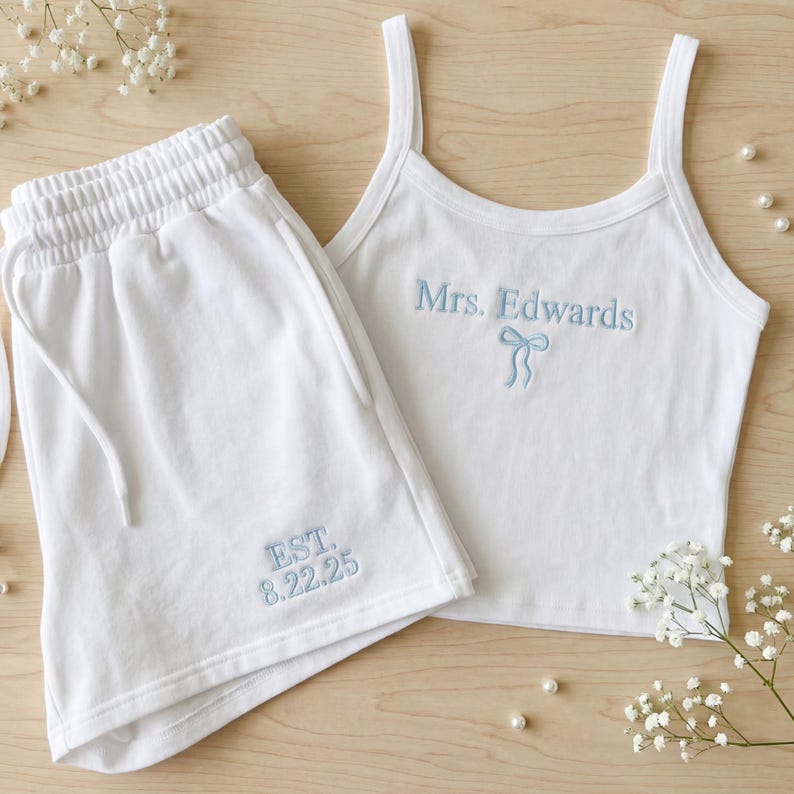 May include: A white two-piece loungewear set. The set includes a tank top and shorts. The tank top has thin straps and light blue embroidered text that reads "Mrs. Edwards" with a bow. The shorts have a drawstring and light blue embroidered text that reads "EST. 8.22.25".