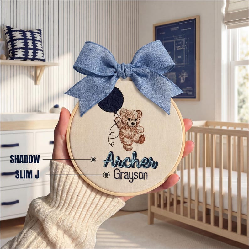May include: Embroidered nursery hoop art featuring a teddy bear holding a balloon, with the name "Archer Grayson" in blue script. A large blue bow tops the hoop. The design is set against a cream-colored fabric, framed by a wooden hoop.