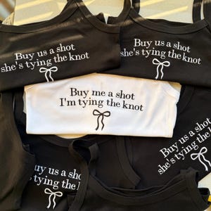 May include: Black and white tank tops and a onesie with the phrases "Buy us a shot she's tying the knot" and "Buy me a shot I'm tying the knot" printed in white. Each top features a bow graphic. Ideal for a bachelorette party.