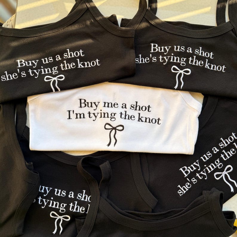 May include: Black and white tank tops and a onesie with the phrases "Buy us a shot she's tying the knot" and "Buy me a shot I'm tying the knot" printed in white. Each top features a bow graphic. Ideal for a bachelorette party.
