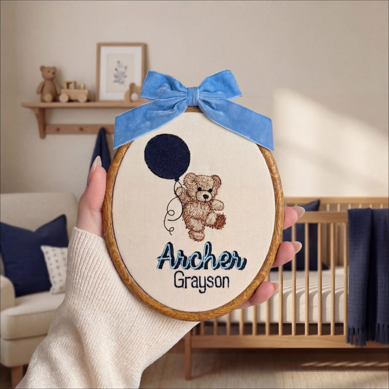 May include: Embroidered nursery hoop art featuring a teddy bear holding a navy blue balloon. The name "Archer Grayson" is stitched in blue thread. A blue velvet bow is attached to the top of the wooden hoop, creating a charming baby room decoration.