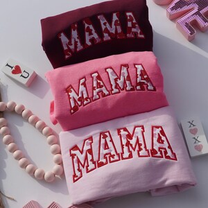 May include: Three sweatshirts in shades of pink and burgundy are stacked. Each sweatshirt has the word "MAMA" embroidered in white with red heart-shaped accents. Decorative wooden beads and small signs with hearts are also visible.