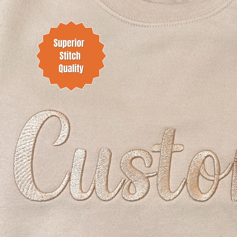 May include: Close-up of a beige sweatshirt with the embroidered word "Custom" in a cursive font. An orange burst graphic above the word reads "Superior Stitch Quality."