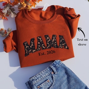 May include: An orange sweatshirt with the word "MAMA" in leopard print lettering and the text "Est. 2026". The sweatshirt is paired with blue denim jeans. The sleeve has text on it.