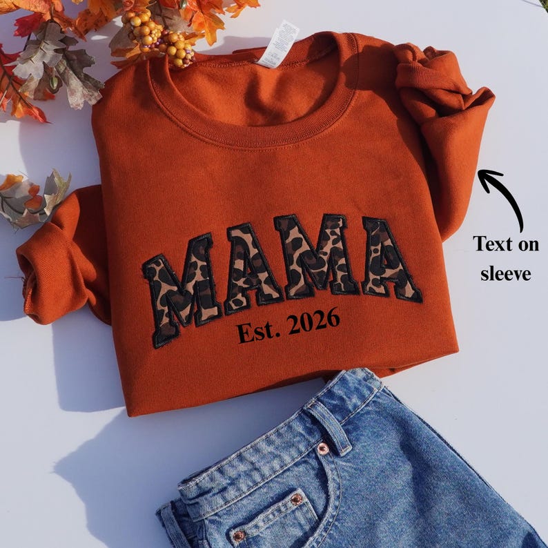 May include: An orange sweatshirt with the word "MAMA" in leopard print lettering and the text "Est. 2026". The sweatshirt is paired with blue denim jeans. The sleeve has text on it.