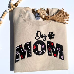 May include: A cream-coloured sweatshirt featuring the embroidered text "Dog MOM". The word "Dog" is in cursive, and "MOM" is in a block letter style with paw prints and bones. A paw print graphic is above the "MOM".