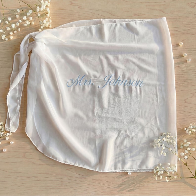 K&ouml;nnte beinhalten: Ein wei&szlig;er, transparenter Wickelrock mit dem gestickten Text Mrs. Johnson in hellblauer Schreibschrift. Das Kleidungsst&uuml;ck verf&uuml;gt &uuml;ber einen Bindeg&uuml;rtel auf der linken Seite und besteht aus leichtem, halbtransparentem Stoff mit weichen Falten.
