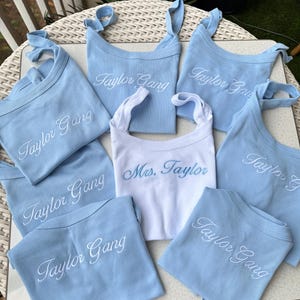 May include: A collection of baby bibs in light blue and white. The light blue bibs are embroidered with "Taylor Gang" in white script. One white bib is embroidered with "Mrs. Taylor" in blue script.