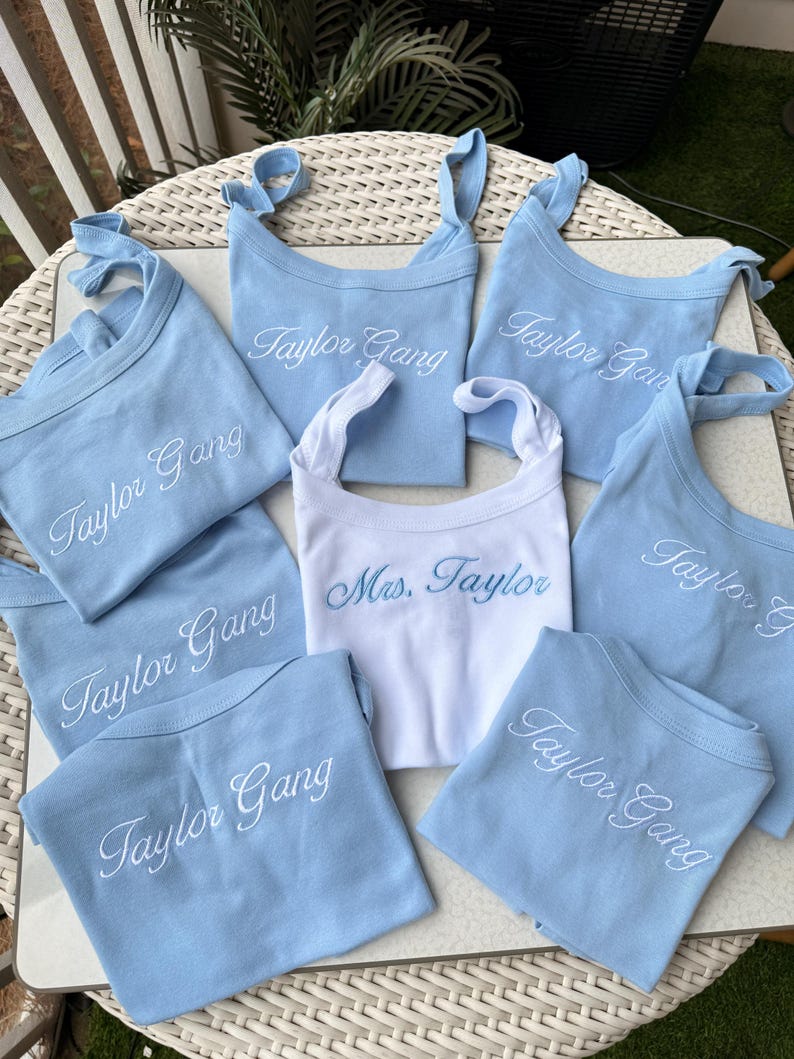 May include: A collection of baby bibs in light blue and white. The light blue bibs are embroidered with "Taylor Gang" in white script. One white bib is embroidered with "Mrs. Taylor" in blue script.