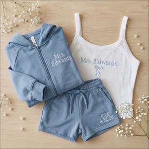 May include: A matching set of loungewear in light blue and white. The set includes a zip-up hoodie, shorts, and a tank top. The words "Mrs. Edwards" are embroidered on the hoodie and tank top. The shorts have "EST. 8.22.25" embroidered on them.