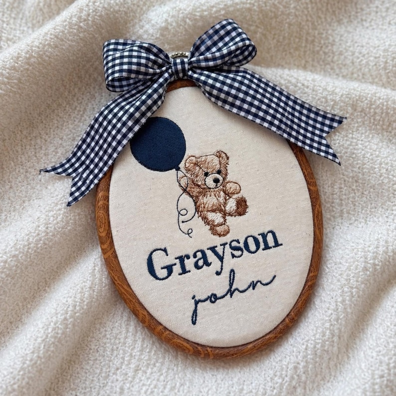 May include: An oval-shaped embroidered nursery hoop art featuring a teddy bear holding a navy blue balloon. The design includes the name "Grayson John" in navy blue script. A gingham bow in black and white adorns the top, adding a decorative touch.