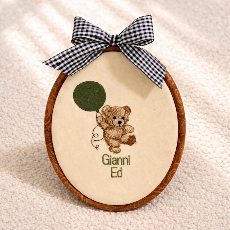 May include: Oval-shaped embroidered artwork featuring a brown teddy bear holding a green balloon. The artwork is framed in a wooden hoop and topped with a black and white checkered bow. The name "Gianni Ed" is embroidered below the bear.