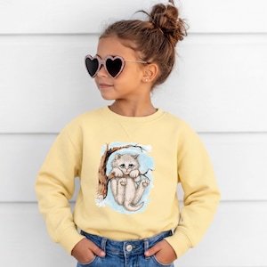 May include: A light yellow sweatshirt featuring a graphic of a fluffy kitten hanging from a tree branch. The kitten has blue eyes and is set against a light blue background. The sweatshirt is long-sleeved and made of a soft material.