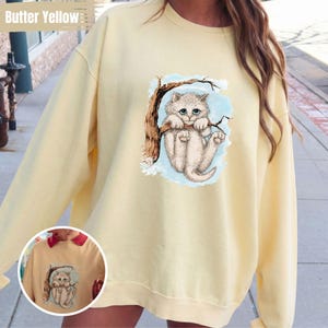 May include: Butter yellow sweatshirt featuring a whimsical illustration of a fluffy white cat clinging to a tree branch. The cat has blue eyes and is set against a light blue backdrop. The words "Butter Yellow" are printed at the top.