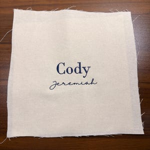 May include: A square, off-white fabric patch with the name "Cody" embroidered in navy blue, above the name "jeremiah" in a cursive font. The edges of the fabric are unfinished, with visible threads.
