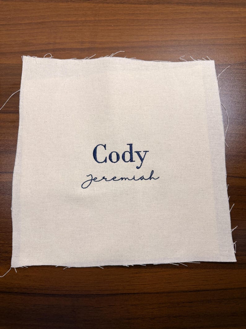 May include: A square, off-white fabric patch with the name "Cody" embroidered in navy blue, above the name "jeremiah" in a cursive font. The edges of the fabric are unfinished, with visible threads.