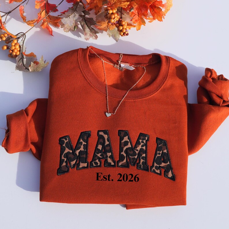 May include: An orange sweatshirt with the word "MAMA" in a leopard print design, with "Est. 2026" underneath. A silver necklace with a heart pendant is draped over the sweatshirt. Autumn leaves and berries are in the background.