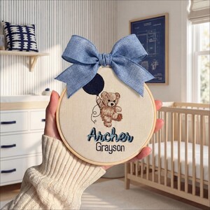 May include: A round, cream-colored embroidery hoop featuring a brown teddy bear holding a black balloon, with the names "Archer Grayson" embroidered in blue. A large blue bow is attached to the top. The hoop is held in front of a nursery setting.
