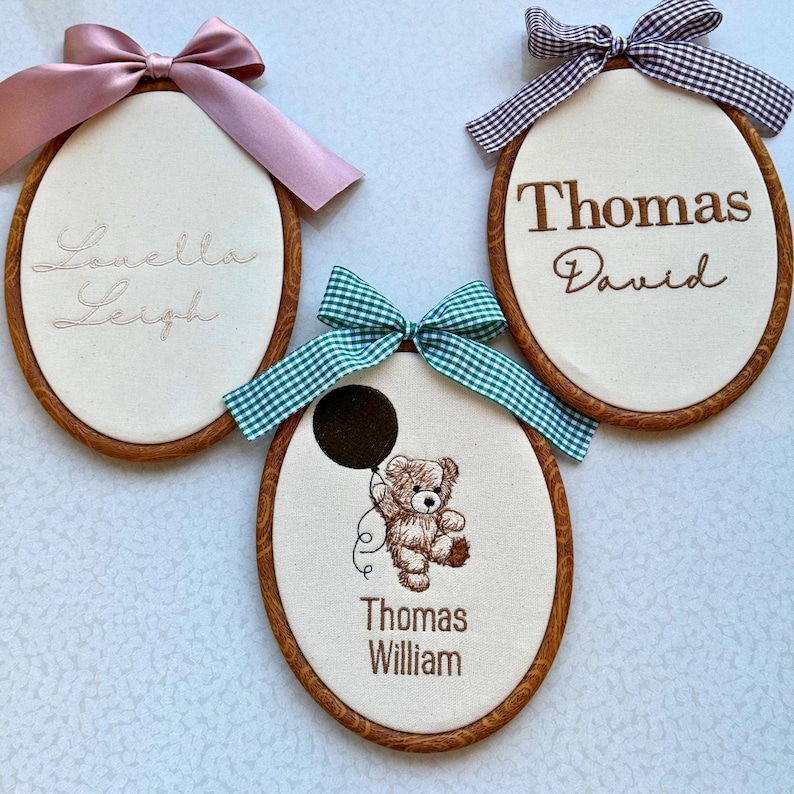 May include: Three oval-shaped embroidered name plaques with wooden frames and bows. One has the name "Louella Leigh" in cursive, another says "Thomas David", and the last one features a teddy bear holding a balloon and the name "Thomas William".