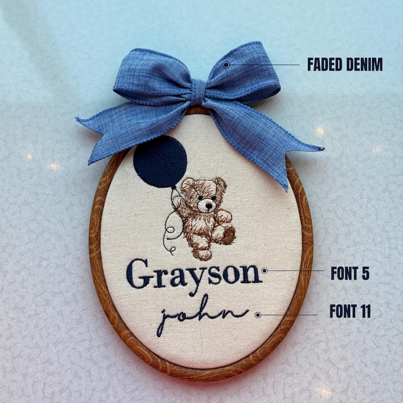 May include: An embroidered oval-shaped wall hanging with a faded denim bow. The design features a teddy bear holding a navy blue balloon, with the name "Grayson" and the name "john" embroidered below. The wooden frame adds a rustic touch.
