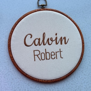 May include: A round, wooden embroidery hoop with the names "Calvin" and "Robert" embroidered in brown thread on a cream-colored fabric. The hoop has a metal hanging loop at the top.