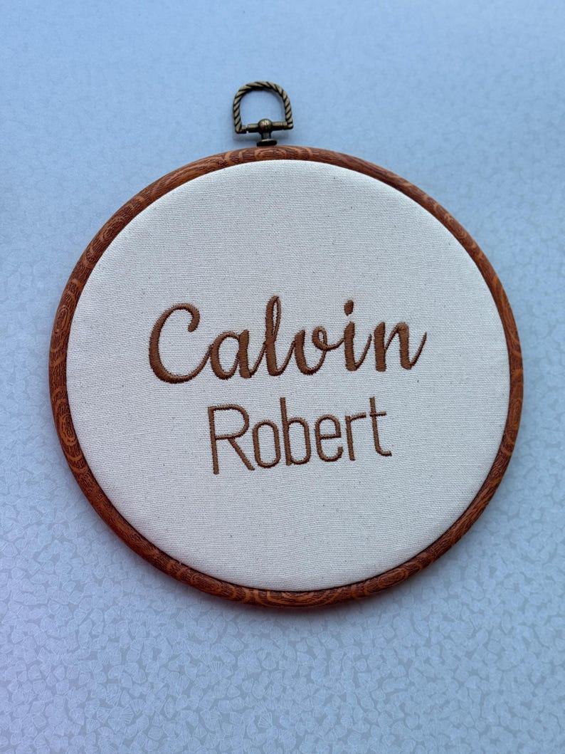 May include: A round, wooden embroidery hoop with the names "Calvin" and "Robert" embroidered in brown thread on a cream-colored fabric. The hoop has a metal hanging loop at the top.