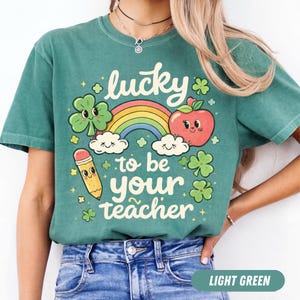 Puede incluir: Camiseta verde claro con un gráfico colorido. El gráfico incluye un arcoíris, una manzana, un lápiz, tréboles y el texto "lucky to be your teacher". La camiseta está hecha de un material suave.