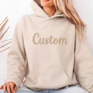 May include: A beige hoodie with a front pocket and a hood. The word "Custom" is embroidered in a cursive font across the chest. The hoodie is a neutral colour.