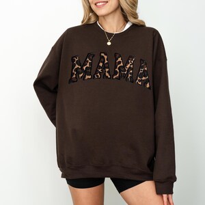 May include: A brown sweatshirt with the word "MAMA" in a leopard print design across the chest. The sweatshirt has a crew neck and long sleeves. The model is wearing black shorts.