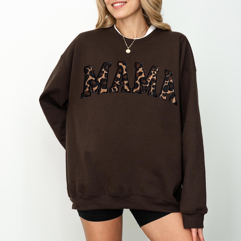 May include: A brown sweatshirt with the word "MAMA" in a leopard print design across the chest. The sweatshirt has a crew neck and long sleeves. The model is wearing black shorts.