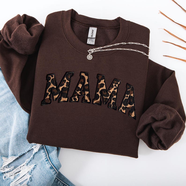 May include: A brown sweatshirt with the word "MAMA" embroidered in a leopard print design. A silver necklace with a sunburst pendant is draped across the sweatshirt. The sweatshirt is folded on top of a pair of blue jeans.