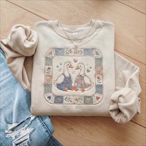 May include: Cream-colored sweatshirt featuring an embroidered design of two geese facing each other, surrounded by a square border with hearts and floral elements. A delicate gold necklace rests on the sweatshirt. The sweatshirt is paired with blue jeans.