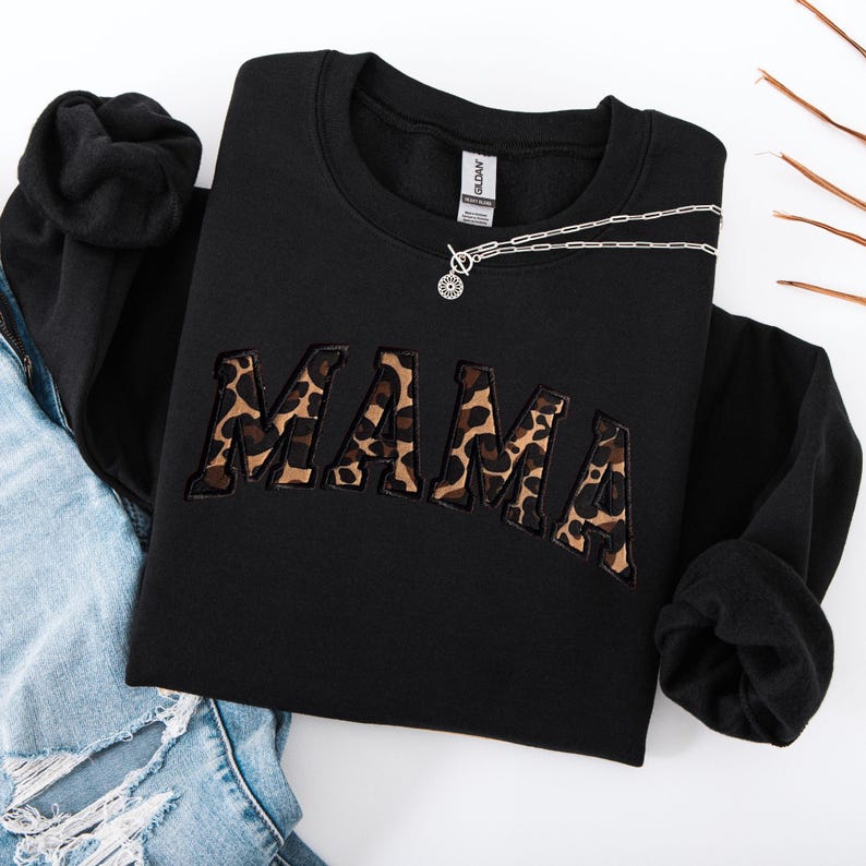 May include: Black crewneck sweatshirt with the word "MAMA" embroidered in a leopard print design. A silver necklace with a circular pendant rests on the sweatshirt. The sweatshirt is folded on top of a pair of blue jeans.