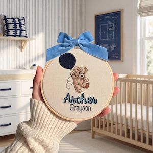 May include: Embroidered nursery hoop art featuring a teddy bear holding a dark blue balloon, with the names "Archer Grayson" in blue script. The artwork is framed in a wooden hoop and adorned with a light blue velvet bow.