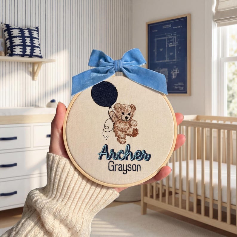 May include: Embroidered nursery hoop art featuring a teddy bear holding a dark blue balloon, with the names "Archer Grayson" in blue script. The artwork is framed in a wooden hoop and adorned with a light blue velvet bow.
