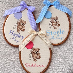 May include: Three embroidered baby name plaques. Each features a teddy bear holding a balloon, with a name and a bow. The plaques have a wooden frame and are decorated with blue, pink, and cream ribbons.