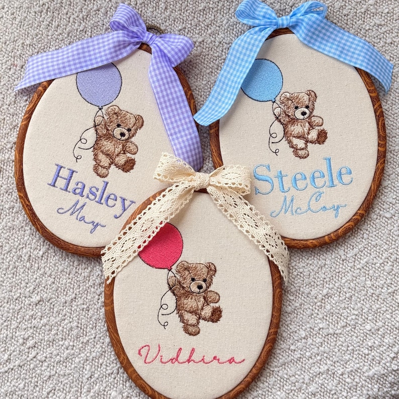 May include: Three embroidered baby name plaques. Each features a teddy bear holding a balloon, with a name and a bow. The plaques have a wooden frame and are decorated with blue, pink, and cream ribbons.