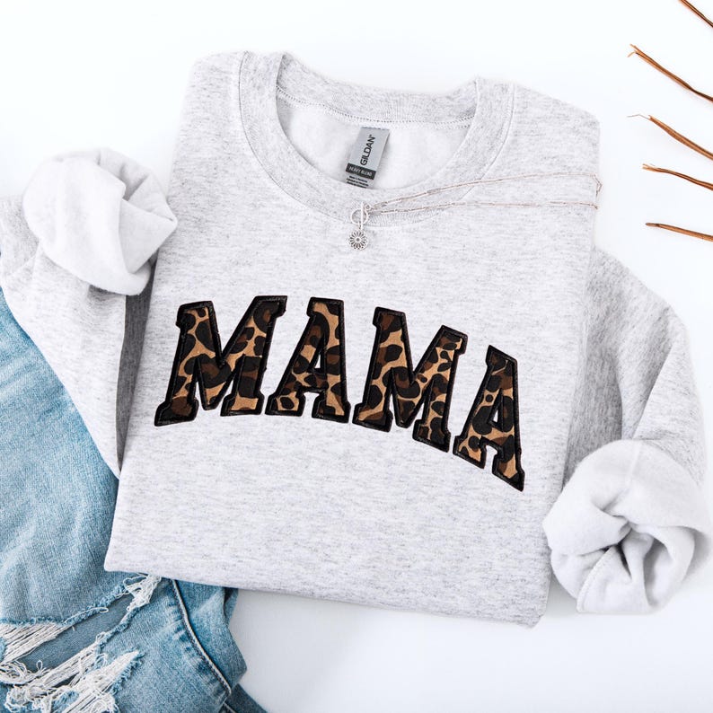 May include: A light gray sweatshirt with the word "MAMA" in a leopard print design. The sweatshirt has long sleeves and a crew neck. A silver necklace with a small pendant is draped across the front. The sweatshirt is folded on top of blue jeans.