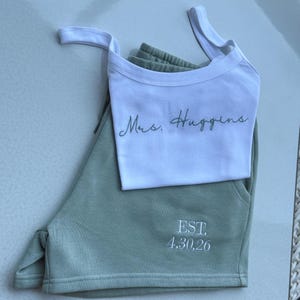 May include: A white tank top and sage green shorts set. The tank top has the text "Mrs. Huggins" in a script font. The shorts have the text "EST. 4.30.26" embroidered in white. The set is made of a soft material.