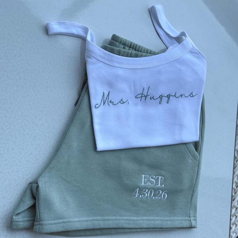May include: A white tank top and sage green shorts set. The tank top has the text "Mrs. Huggins" in a script font. The shorts have the text "EST. 4.30.26" embroidered in white. The set is made of a soft material.