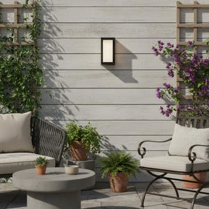 May include: An outdoor patio setting featuring a black rectangular wall-mounted light. The light is fixed to a light grey wooden wall. The scene includes a round concrete table, two chairs, and potted plants. The chairs have cream-coloured cushions.