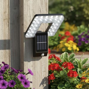 May include: A black solar-powered outdoor light fixture with a motion sensor. The light has a rectangular solar panel and a square light array with multiple bright white LED lights. It is mounted on a wooden post, with colorful flowers in the background.