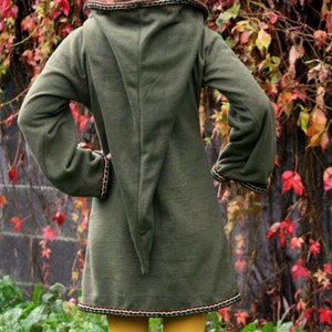 Puede incluir: Una túnica larga de capucha verde con una cola larga y puntiaguda. La túnica tiene un ribete marrón, verde y blanco alrededor de la capucha y el dobladillo. La persona que lleva la túnica está de pie frente a una pared de enredaderas verdes con hojas rojas.