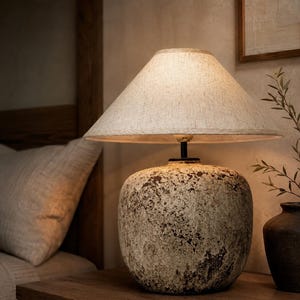 May include: A table lamp featuring a rounded, textured ceramic base with a mottled beige and dark brown finish. The lamp is topped with a conical, off-white fabric lampshade that emits a soft, warm glow, illuminating the surrounding area.