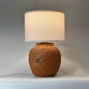 Puede incluir: Una lámpara de mesa rústica con una base de cerámica texturizada de color terracota y una pantalla de color crema. La lámpara tiene una base redonda en forma de jarrón y una pantalla cilíndrica.